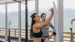 Female fitness coach writes on whiteboard as a male athlete watches in a gym.